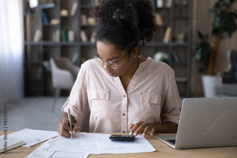 Serious young biracial woman sit at desk manage budget calculate on machine pay bills taxes online on laptop. Focused African American female count expenses expenditures on calculator. Save concept.
